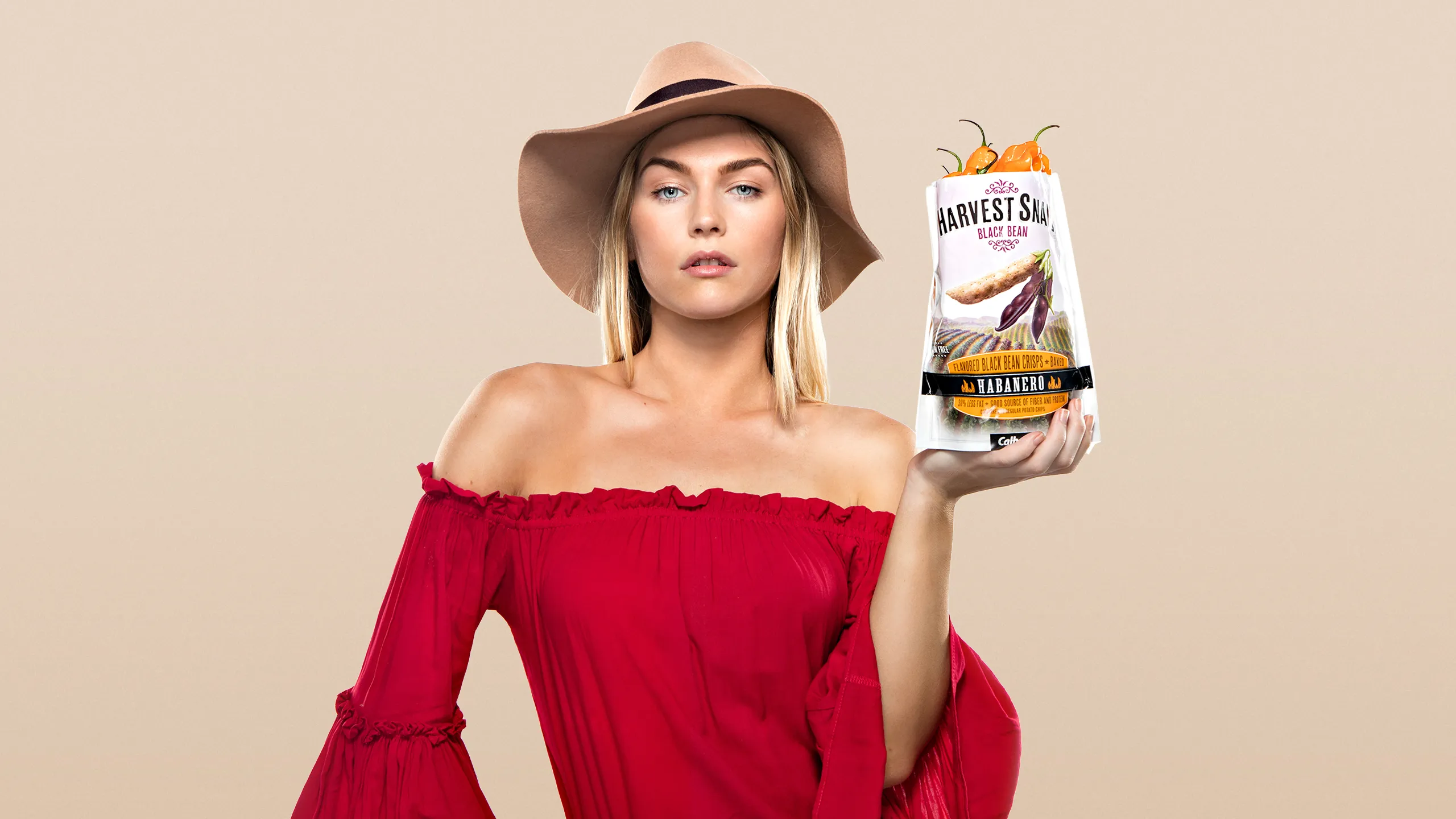 Woman in a red off-shoulder top holding a bag of Harvest Snaps Habanero Black Bean Crisps
